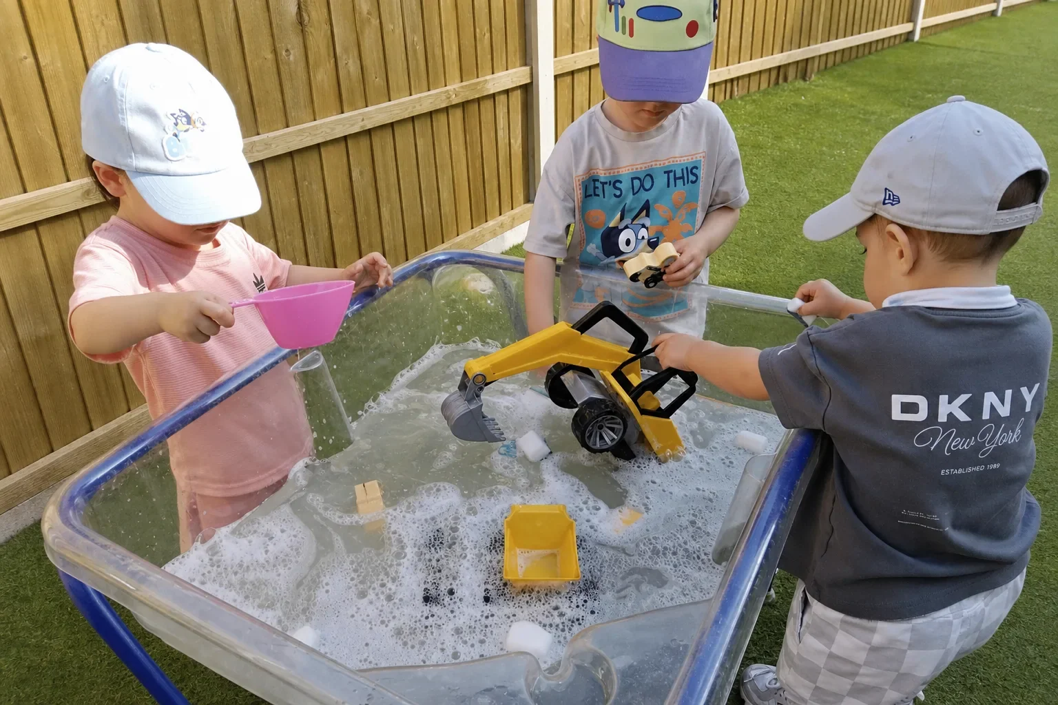 Three young children playing together with vehicles, cups and bubbly water at an outdoor water table